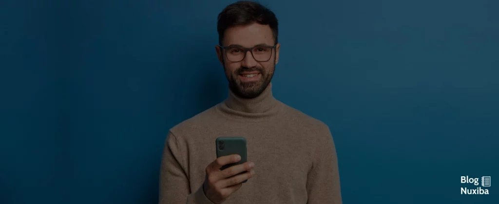 Hombre con lentes y barba, joven, sonriendo frontal a cámara, sosteniendo móvil en la mano derecha sobre fono azul.