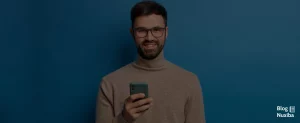 Hombre con lentes y barba, joven, sonriendo frontal a cámara, sosteniendo móvil en la mano derecha sobre fono azul.
